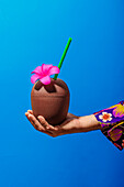 Körperteil Mann in lebhaftem Hemd mit ornamentalem Blumenmuster hält Kokosnusscocktail mit Strohhalm und Blumendekoration auf blauem Hintergrund