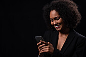 Positive Hispanic young female in outfit smiling and browsing mobile phone while standing on black background