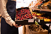 Side view of cropped unrecognizable male seller showing box of fresh ripe plums standing near assorted fruits and vegetables in supermarket