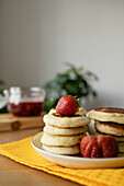Köstliche süße Erdbeeren auf gestapelten Pfannkuchen auf Teller über Tischtuch auf Holztisch mit Erdbeermarmelade in hellem Raum