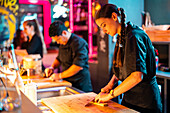 Side view of young female chef with braids in black uniform with focus cutting ingredient with knife on chopping board while preparing meal at illuminated counter in bistro