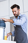 From below of adult male chef in apron looking down while standing at kitchen table with juice squeezer during healthy breakfast process in daylight