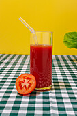 Transparent glass of fresh tomato juice with glass straw placed on checkered tablecloth against yellow background