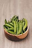 A wooden bowl filled with ripe green peas is set against a textured wooden backdrop, presenting a natural and rustic image.