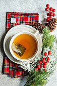 Top view of Christmas cup of tea placed on red napkin near holly and pine cones on gray table