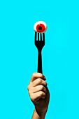 Hand of crop anonymous person holding eyeball with red iris on fork isolated on vivid blue backdrop