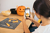 Junge hispanische Frau schaut von oben auf den Bildschirm ihres Mobiltelefons, während sie am Tisch sitzt und ein Foto von einem verschwommenen Jack o Lantern-Kürbis am Tag macht