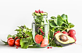 Green salad in jar with ingredients: green lettuce leaves, tomato, strawberry, avocado, cucumber and pomegranate seeds at white background. Healthy to go food with vegetables and fruits. Front view.