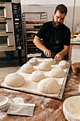 Concentrated serious mature male chef scoring uncooked dough with knife on large baking tray at table while making bread in bakery