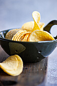 Closeup of stacked pieces of delicious crispy yellow potato chips in black bowl over wooden table in bright room against blurred background
