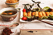 An Asian dining setup featuring vietnamese rolls on a plate, a bowl of soup, rice, and chopsticks.