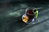 Blick von oben auf ein klares Glas Tee mit einer Zitronenscheibe und einem grünen Teesieb auf einem strukturierten dunklen Hintergrund
