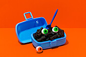 Horror-Lunch mit schwarzer Nudelgabel und Augen in Lunchbox mit Gabel auf orangem Hintergrund