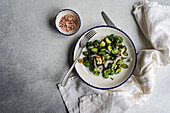 Draufsicht auf einen Gurkensalat mit roten Zwiebeln, Oliven und würzigem Käse, serviert auf einem weißen Teller mit Gabel und Messer neben einer Serviette auf einem grauen Tisch im Tageslicht