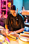 Confident young female chef with long braids in black uniform and gloves standing while placing freshly cooked sushi on tray at kitchen at restaurant