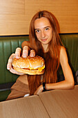 Young woman with red hair and piercing sitting at table and looking at camera while showing hamburger in restaurant
