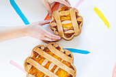 Abgeschnittene, nicht erkennbare Hände arrangieren italienisches Ostergebäck Pastiera napoletana auf einer weißen Fläche mit bunten Pinselstrichen