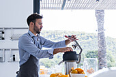 Side view of focused adult male chef in apron looking down while standing at kitchen table and putting orange into juice squeezer during healthy breakfast process in daylight