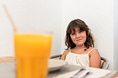 Portrait of smiling cute little girl in casual clothes sitting on chair with fruit on table and looking at camera against white wall in restaurant
