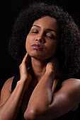 Hispanic young female with curly hair touching neck with closed eyes while standing on black background