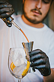 Concentrated young professional barman pouring orange drink in jigger while preparing refreshing cocktail and standing at counter in bar