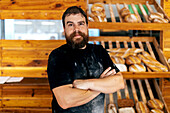 Portrait of confident positive bearded mature man with arms crossed standing against wooden shelves with freshly baked breads in bakery
