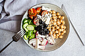 Schale mit gesundem Salat und Gemüse mit Kichererbsen, Sesam und Oliven mit Besteck und Stoff auf einem hellgrauen Tisch von oben