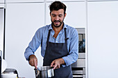 Smiling bearded adult male chef in apron looking down while standing in kitchen and mixing ingredients in saucepan during breakfast preparation in modern kitchen in daylight