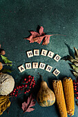 Komposition aus reifem, gesammeltem Kürbis und Maiskolben, arrangiert mit Spielwort-Holzfliesen mit der Aufschrift "Hello Autumn" auf grünem Hintergrund