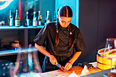 Focused young female chef in uniform and with long braided hair cutting meat with knife on counter while preparing meal in bistro