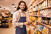 Portrait of happy ethnic male worker with long curly hair in casual clothes standing near shelves looking at camera smiling while working on tablet in modern lighted grocery shop