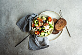 Draufsicht auf eine Keramikschale mit leckerem, gesundem Salat, Gemüse und Brot mit Gurken- und Tomatenscheiben, Besteck und Stoff auf einem grauen Tisch