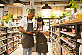 Young African American male and female sales staff in apron standing in between cupboards with grocery items and looking at screen of tablet while browsing