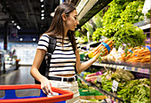 Side view of young female customer in casual clothes with shopping basket choosing fresh carrots while standing near shelves in supermarket