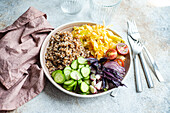 High angle of bowl with delicious buckwheat containing sliced fresh basil plant with cabbage and cucumber with cutlery and glasses placed on gray table with napkin