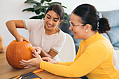 Happy female friends looking down while sitting at wooden table with Halloween tools and carving eye in daylight