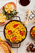 Draufsicht auf eine traditionelle spanische Paella in einer Pfanne, serviert mit Brot, Wein und Beilagen auf einem gemusterten Tischtuch