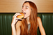 Young woman with red hair and piercing with closed eyes sitting at table and enjoy eating hamburger in restaurant