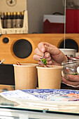 Closeup of Crop anonymous person's hand adding fresh green garnish to soup containers against blurred background