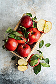 Top view of whole fresh red ripe organic delicious apples with green leaves filled in wooden box with half cut pieces and placed on gray surface in daylight