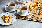 Hoher Winkel von Tellern mit Stücken von Apfelkuchen und Tassen Tee neben Messer, Gabel und Serviette auf einem rustikalen Tablett vor einem grauen Tisch
