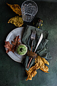 Top view of autumnal table setting with napkin, knife and fork place on plate near colorful leaves, smalls pumpkins and empty glass against dark surface