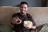 Inhalt: Junger ethnischer Afroamerikaner in Freizeitkleidung sitzt mit einer Schüssel Popcorn und schaltet den Fernseher mit schwarzem Bildschirm ein, während er sich zu Hause auf einem bequemen Sofa ausruht
