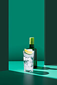 Eine minimalistische Komposition mit einer Gin-Flasche hinter einem Glas Tonic Water, das mit einer Limettenscheibe verziert ist, alles vor einem zweifarbigen grünen Hintergrund
