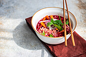 High angle of bowl with beetroot soup with onion, coriander and noodles in Asian style served on bowl and chopsticks on napkin against gray surface in daylight