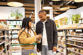 Cheerful young multiethnic couple in casual wear choosing goods in supermarket together while shopping with grocery cart during sale