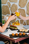 Unrecognizable crop people sitting at table in restaurant with yummy delicious dishes served along with empty plates and saying cheers with drink glasses in daylight