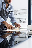 Anonymous crop bearded male with folded sleeves and apron standing at reflecting surface kitchen table while cutting cheese against blurred background in daylight