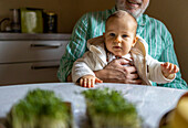 Crop anonymous senior grandfather in casual clothes holding adorable baby boy while sitting by table at home