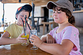 Cute brother and sister wearing casuals and caps toasting fresh fruit juices while sitting together at table in restaurant during summer vacation